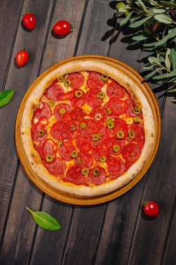 Taze pişmiş pepperonili pizza ahşap bir masanın üzerinde duruyor, yeşil zeytin ve vişneli domateslerden oluşan canlı bir tepsi sergileniyor. Pizzanın etrafında taze fesleğen yaprakları ve dağınık domatesler var..