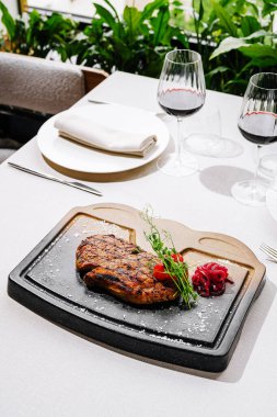 Güzel bir şekilde sunulmuş ızgara biftek sıcak bir taşın üzerine konur ve yanında süslemeler de vardır. Yemek masası, arka planda şarap kadehleri ve yeşillik ile zarifçe ayarlanmış..