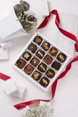 Çok güzel düzenlenmiş bir kutu çikolata beyaz bir yüzeyde duruyor. Karmaşık malzemeler içeriyor. Kurutulmuş çiçekler ve kurdele gibi dekoratif elementlerle çevrili..