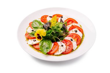 Canlı bir caprese salatası, art arda domates ve mozzarella dilimleri sergiliyor. Taze fesleğen ve yenilebilir çiçeklerle süslenmiş. Beyaz bir tabakta ustalıkla sergileniyor..
