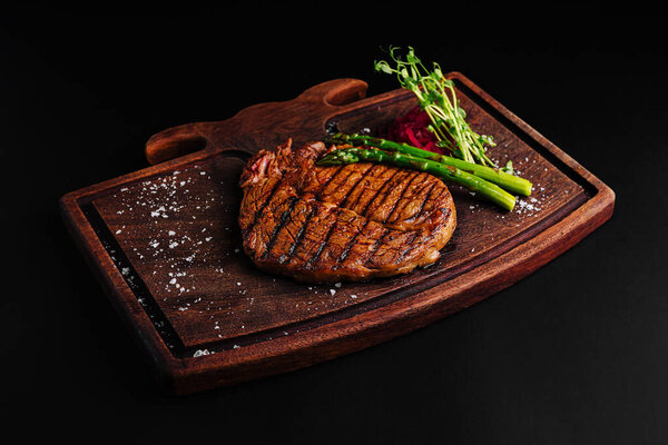 A perfectly grilled steak rests on a wooden cutting board, accompanied by fresh asparagus and herbs. The scene highlights rich textures and colors, creating an appetizing presentation.