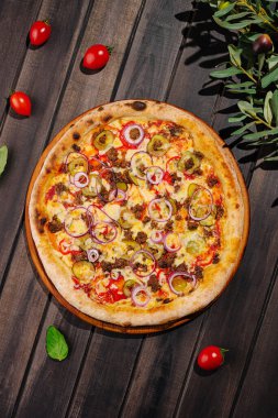 Yuvarlak bir pizza, zeytin, domates, soğan ve otlar gibi canlı malzemelerle dolu ahşap bir yüzeyde sergileniyor, ağız sulandıran bir yemek sunulmaya hazır..