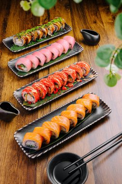 Renkli suşi dürümleri siyah tepsilerde sıralanır, çeşitli malzemeler ve stiller sergilenir. Sunuma soya sosu ve çubukları eşlik ederek yemek deneyimini artırır.