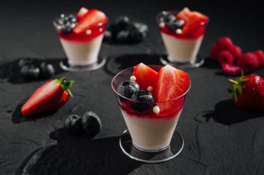 Taze çilek, yabanmersini ve ahududu soslu kremalı panna cotta ile dolu üç zarif fincan. Koyu arkaplan, dutların canlı renklerini vurguluyor..