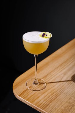 Ferahlatıcı bir kokteyl narin beyaz bir köpük ve canlı bir çiçekle kaplı uzun bir bardakta zarif bir şekilde oturur. Ahşap masa, karanlık bir arkaplan karşısında sunuma sıcaklık katar..