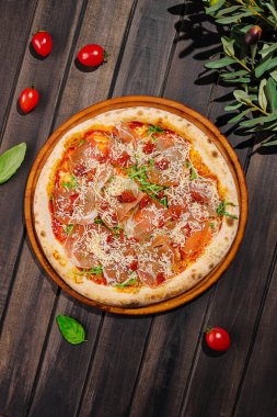 Tahtaya konmuş lezzetli bir pizza domates sosu, prosciutto ve peynir içerir. Etrafında taze fesleğen yaprakları ve kiraz domatesleri var. Canlı bir mutfak sunumu yapıyorlar..
