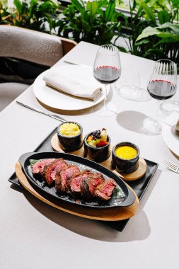 Nefis bir ızgara biftek, arka planda yeşillik olan zarif bir restoranda canlı yan yemeklerle birlikte siyah bir taş tabakta duruyor. Güzel bir yemek deneyimi için ideal..