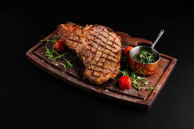 Güzel pişirilmiş bir biftek ahşap bir tepside sunulur, taze otlar ve kiraz domatesleriyle süslenir. Küçük bir kase sos yemeği tamamlıyor, gurme mutfağı deneyimi sergiliyor..