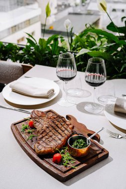Güzel hazırlanmış bir bifteğe ahşap bir tahtada ızgara kesilmiş ve yanında zengin yeşil otlar ve vişneli domatesler eşlik ediyor. İki kadeh kırmızı şarap serin bir ortama zarafet katar..
