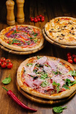 Üç çeşit pizza, ahşap tahtalarda güzelce düzenlenmiştir. Üstlerinde sebzeler, peynir, pişirilmiş etler ve taze otlar var. Sergiye kiraz domates ve baharatlar eşlik ediyor.