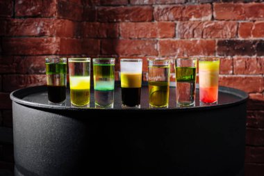 Parlak ve renkli içeceklerle dolu yedi shot bardağı şık bir tezgahın üzerinde duruyor. Çeşitli renkler ışığı yansıtıyor ve enerjik bir atmosfer yaratıyor, gece için mükemmel..