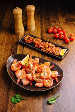 Çok güzel düzenlenmiş bir tabakta sulu karides otlar ve limonla süslenir. Izgara deniz ürünleri ve taze kiraz domatesleri ahşap bir masada canlı sunumu tamamlar..