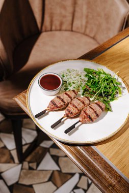 Lezzetli ızgara et şişleri beyaz bir tabakta, yanında taze salata ve küçük bir kase sosla birlikte sıcak bir yemek alanında ahşap bir masa üzerinde hazırlanır..