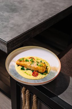 Çağdaş bir yemek masasında sergilenen güzel bir tabak somon filetosu kremalı sosun üzerinde otlar ve vişneli domateslerle duruyor. Ayarlar şık ve zarif bir atmosferi yansıtıyor..