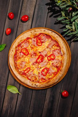 Taze pişmiş bir pizza, üzerinde kırmızı biber ve lezzetli jambon bulunan altın bir kabuk içerir. Etrafında kiraz domatesleri ve ahşap bir masada taze fesleğen yaprakları var..