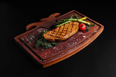 Mükemmel ızgara bir biftek ahşap bir kesme tahtasının üzerinde sergilenir. Taze kuşkonmaz ve vişneli domatesle birlikte, bu yemek iştah açıcı ve gurme yemek deneyimi için iyi hazırlanmış görünüyor..