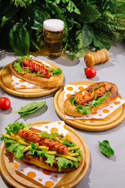 Tahta tabaklar üzerinde taze marul ve domatesle kaplı üç lezzetli sosisli sandviç var. Bir bardak soğuk bira yakınlarda, yeşilliklerle çevrili, rahat bir açık hava yemek deneyimini çağrıştırıyor..