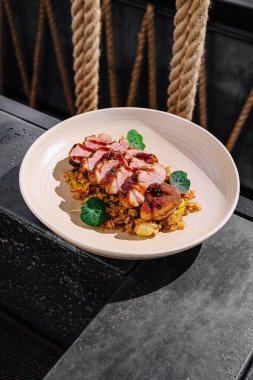 Izgara tavuk dikkatlice dilimlenir ve baharatlı pirincin üzerine konur, yeşillikle süslenir. Tabak, şık bir arkaplan karşısında desenli sofra takımının üzerine konur..