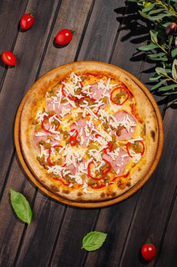 Lezzetli bir pizza ahşap bir masaya konur, üzerine jambon, peynir, biber ve bitkiler konur. Vişneli domates ve taze fesleğen pizzanın etrafını sarar, iştah açıcı görünümünü arttırır..