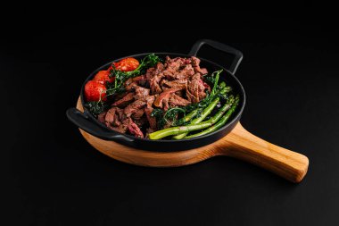 Mükemmel pişirilmiş sığır eti dilimleri, canlı yeşiller ve sulu domateslerle sotelenmiş ahşap bir tabakta servis edilen dumanı tüten bir tabak. 