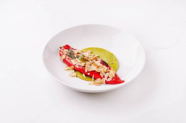 Canlı kırmızı biber, dilimlenmiş bademlerle kaplı dairesel beyaz bir tabakta duruyor ve yeşil sosta servis ediliyor, yaratıcı bir mutfak sunumu sunuyor..