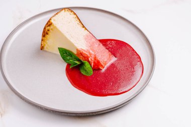 Çökmüş bir cheesecake dilimi zarif bir şekilde yuvarlak bir tabakta sunuluyor. Tatlı, yanında serpiştirilmiş canlı bir ahududu sosu ve garnitür olarak taze bir nane yaprağı içeriyor..