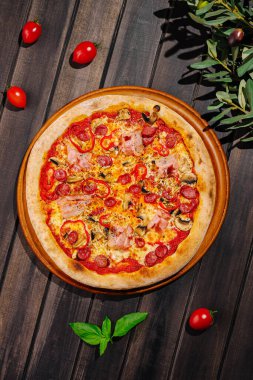 Yeni pişmiş bir pizza ahşap servis tahtasının üzerinde, pepperoni, jambon ve mantar dilimleriyle süslenmiş olarak oturur. Etrafında vişneli domatesler ve taze fesleğenler var. Canlı bir sunum yapıyorlar..