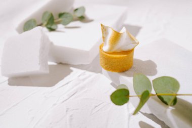 Krema soslu küçük sarı bir tatlı beyaz geometrik şekillerin arasında duruyor. Yeşil okaliptüs yaprakları zarif ve modern tasarımı vurguluyor..