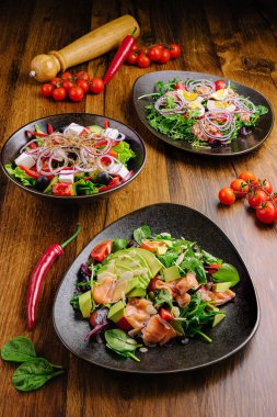 Yaratıcı bir şekilde düzenlenmiş üç salata canlı yeşillikler ve sade ahşap bir masa üzerinde taze malzemeler, kiraz domatesleri ve biberlerle süslenmiş, davetkar bir mutfak sunumu yaratıyor..