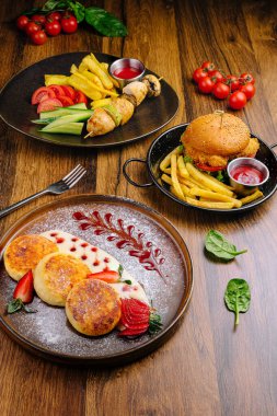 Bir gurme yemek deneyimi, canlı bir gözleme tabağı sunar çilekle süslenmiş, patates kızartmasının yanında lezzetli bir burger, ve ahşap bir masada renkli ızgara şişler..