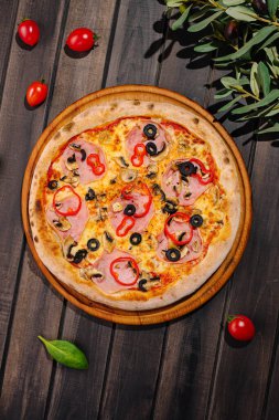 Yuvarlak bir pizza, ahşap bir tahtaya yerleştirilmiş jambon, siyah zeytin ve kırmızı dolma biberlerle süslenir. Tabağın etrafı kiraz domatesleri ve yeşil yapraklarla çevrili..