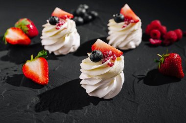 Üzerinde çilek, yaban mersini ve ahududu bulunan üç zarif krem şantili tatlı koyu bir yüzeye dizilir ve renklerin ve dokuların hoş bir kombinasyonunu sergiler..