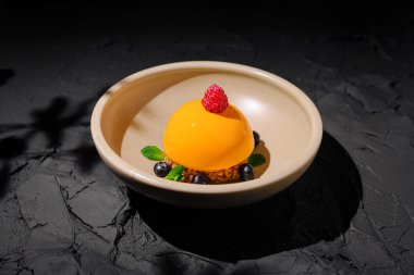 Canlı turuncu bir tatlı bej bir tabağın üzerinde, ahududu ve yaban mersiniyle süslenmiş olarak oturur. Yaradılış, taze nane ile çevrili pürüzsüz bir doku sergiliyor, zarif bir sunum sunuyor..