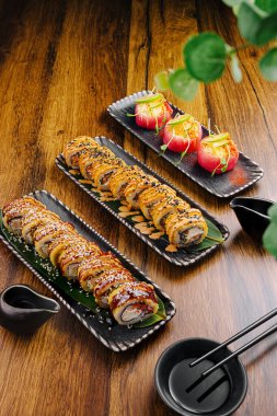 Suşi ruloları ve renkli aperatiflerin yer aldığı üç güzel düzenlenmiş yemek sunuluyor. Ahşap masa, gıdanın canlı renklerini ve dokusunu yükselterek hoş bir atmosfer yaratıyor..