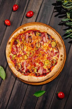 Taze pişmiş bir pizza, lezzetli pastırma, peynir ve turşuyla kaplı ahşap bir tahtanın üzerinde sunulur. Pizzayı çevreleyen kiraz domatesleri ve fesleğen yaprakları canlı bir ortam oluşturuyor..