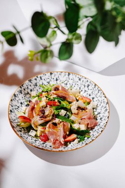 İçinde prosciutto, taze yeşiller, renkli sebzeler ve süslemeler bulunan canlı bir salata. Çiçek desenli bir kasede hafif bir arka planda güzelce sunuluyor..