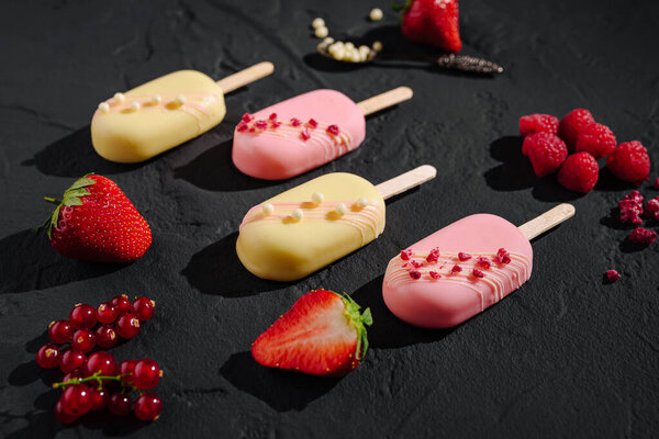 Красочные замороженные лакомства аккуратно расположены рядом со свежей клубникой, малиной и красной смородиной на темной поверхности.