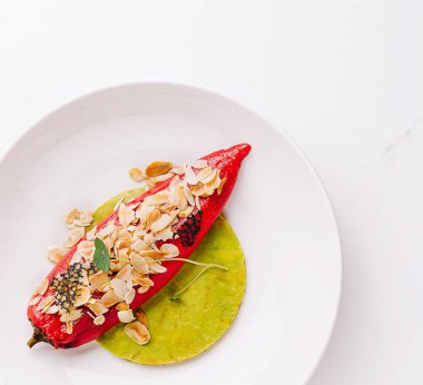 Canlı kırmızı biber yeşil bir tabakta sunuluyor, üzerinde dilimlenmiş badem ve biraz şifalı bitkiler var, yaratıcı bir mutfak sunumu sergileniyor..