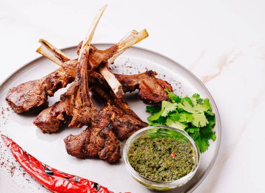 Izgara kuzu pirzolası, canlı bir bitki sosu ve kırmızı biberle birlikte bir tabakta güzelce düzenlenir. Ağız sulandırıcı bir mutfak görüntüsü oluşturur. Her yemek için mükemmel..