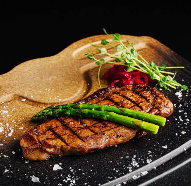 Mükemmel ızgara bir biftek taze kuşkonmaz ve bitkilerle süslenmiş şık bir tabakta oturur. Yemek bir tutam tuz ile sunuluyor, lezzetleri ve dokuları vurguluyor..