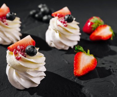 Üzerinde çilek, yaban mersini ve ahududu bulunan üç zarif krem şantili tatlı koyu bir yüzeye dizilir ve renklerin ve dokuların hoş bir kombinasyonunu sergiler..