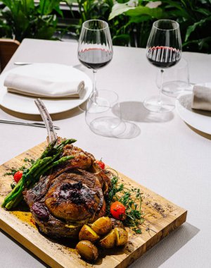 Titizlikle hazırlanmış bir kuzu pirzolası kızarmış patates, kuşkonmaz ve vişneli domatesle servis edilir. Zarif şarap kadehleri yeşilliklerle çevrili taze yemek takımını tamamlıyor..