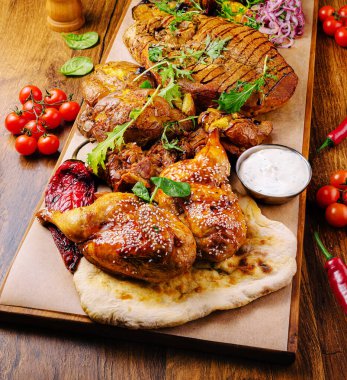 Tavuk ve kuzu eti de dahil olmak üzere canlı bir ızgara ahşap tabakta güzelce düzenlenmiştir. Etin yanında taze bitkiler, renkli süsler ve yanında kremalı sos var..