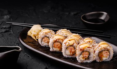 Lezzetli suşi dürümleri küçük bir kase soya sosuyla birlikte koyu ahşap bir tabakta dizilmiş. Wasabi ekmeklerin yanına oturur ve çubuklar zarif bir şekilde yan tarafa yerleştirilir..