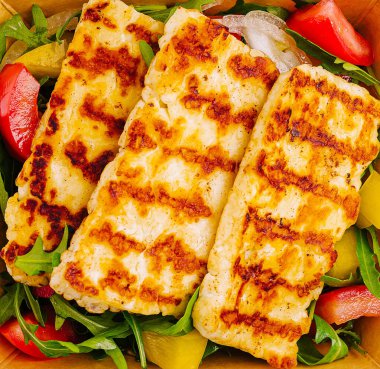 Izgara halloumi peyniri roka ve rengarenk sebzelerden oluşan bir yatakta sergileniyor. Lezzetli ve sağlıklı bir yemek seçeneği sunuyor. Yaz yemeği için mükemmel..