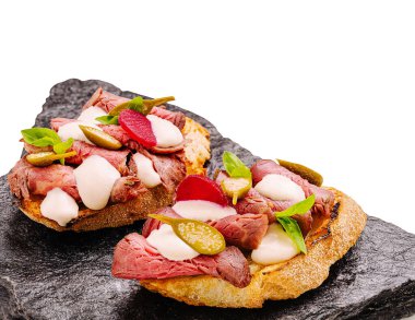 Bir sandviç, turşu ve taze otlar da dahil olmak üzere renkli malzemelerle bezenmiş yumuşak bir biftek içerir, siyah taştan bir tabakta zarif bir şekilde sunulur..