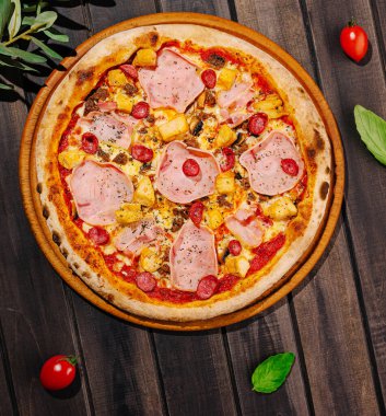 İçinde jambon, ananas ve vişneli domatesler bulunan taze pişmiş bir pizza ahşap bir masada duruyor. Etrafında yeşil yapraklar ve canlı kırmızı domatesler var..