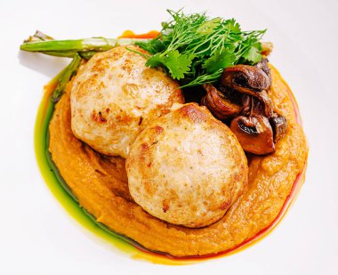 Yumuşak bir sebze püresi üzerine servis edilen yumuşak tavuk köfteleri yanında sotelenmiş mantarlar, kavrulmuş kuşkonmaz ve bir gurme sunumu için zarif bir şekilde kaplanmış taze otlar..