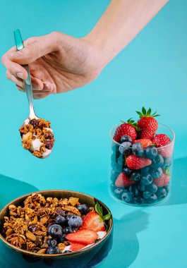 Bir el, canlı turkuaz bir yüzey üzerinde kavanozlar dolusu meyve ile tamamlanmış çilek ve yaban mersinleriyle dolu bir kasenin üzerinde bir kaşık dolusu altın granola tutar..