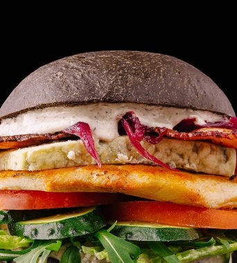 Görsel olarak çarpıcı bir hamburgerde ızgara tavuk, marul ve domates gibi taze sebzeler ve kremalı sosla dolu siyah bir çörek bulunur. Modern sunum katmanlarını ve dokularını vurguluyor.